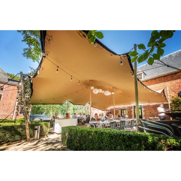 een stretchtent van 10,5x15 meter zit met houten palen vast in de grond. Er staan twee palen in het midden om de gehele tent omhoog te houden. de houten palen zijn vastgezet met touwen en haringen die in de grond zitten.