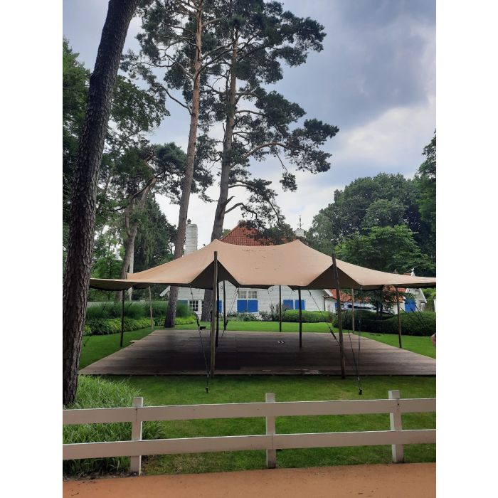 Een stretchtent van 10,5x10,5 meter zit met houten palen vast in de grond. Er staan twee palen in het midden om de gehele tent omhoog te houden. de houten palen zijn vastgezet met touwen en haringen die in de grond zitten.