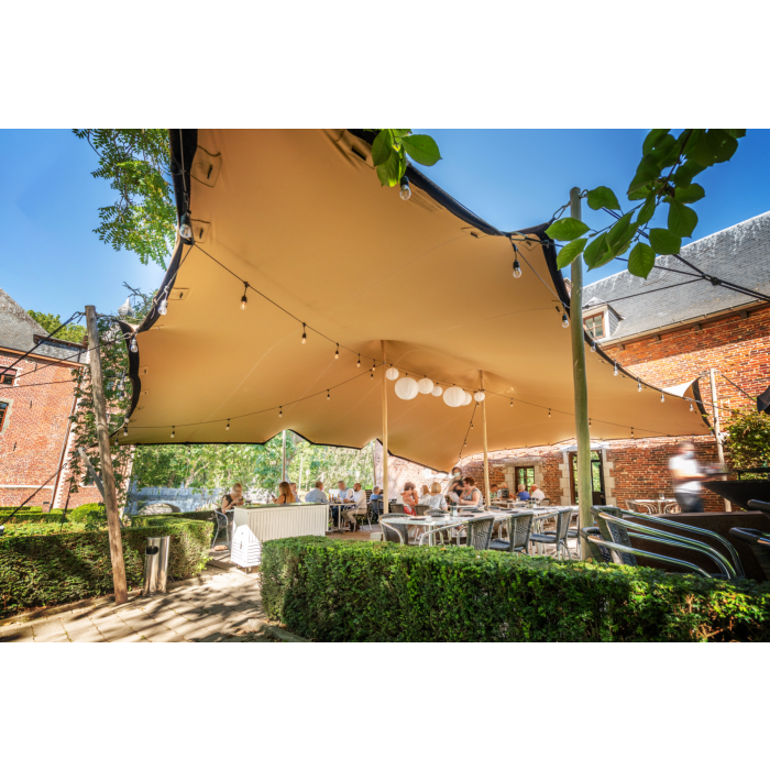een stretchtent van 10,5x15 meter zit met houten palen vast in de grond. Er staan twee palen in het midden om de gehele tent omhoog te houden. de houten palen zijn vastgezet met touwen en haringen die in de grond zitten.